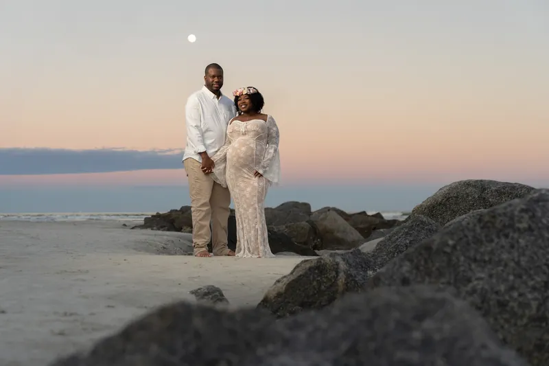 Ashley and JB stand together at sunset on Vilano Beach, with the moon visible above the calm water and rocky shoreline behind