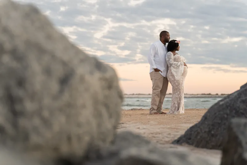 Ashley and JB embrace on the sand at Vilano Beach, framed by large rocks in soft evening light.