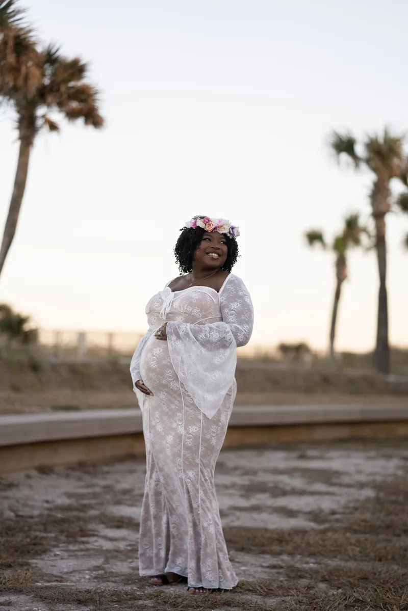 Ashley in white lace dress with flower crown, standing among palm trees at sunset