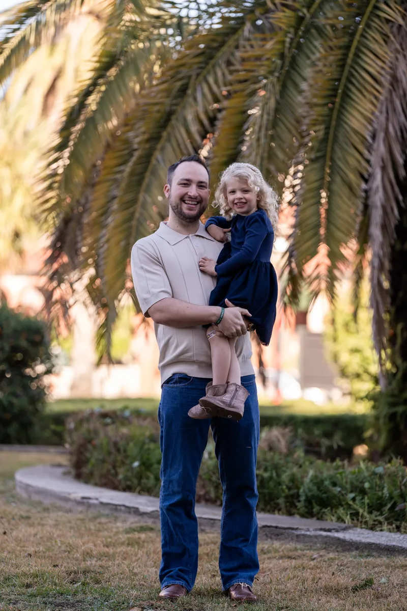 A father holds his young daughter in his arms, both smiling warmly at the camera under beautiful palm trees during golden hour.