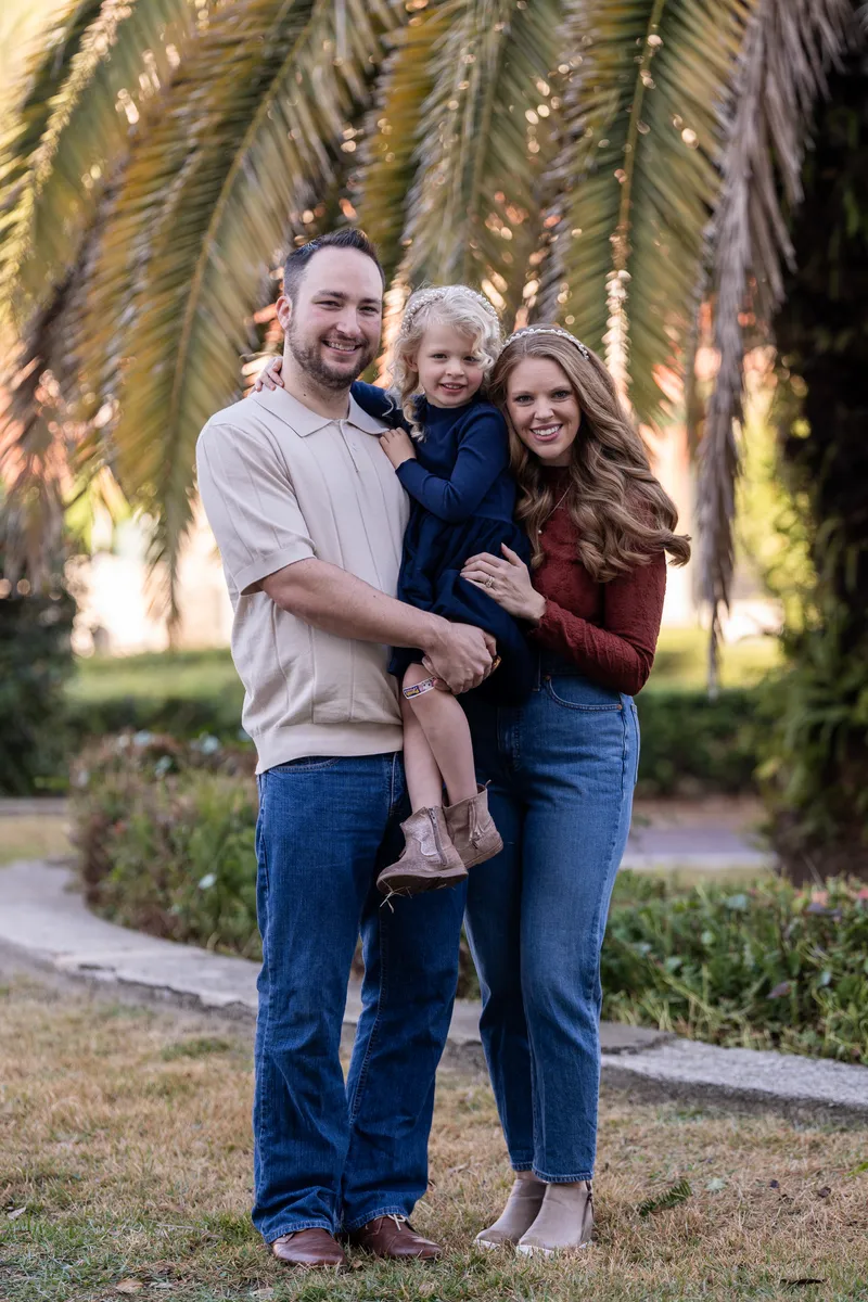 Ashley and Jordan embrace their young daughter in a warm family portrait beneath palm trees during golden hour.