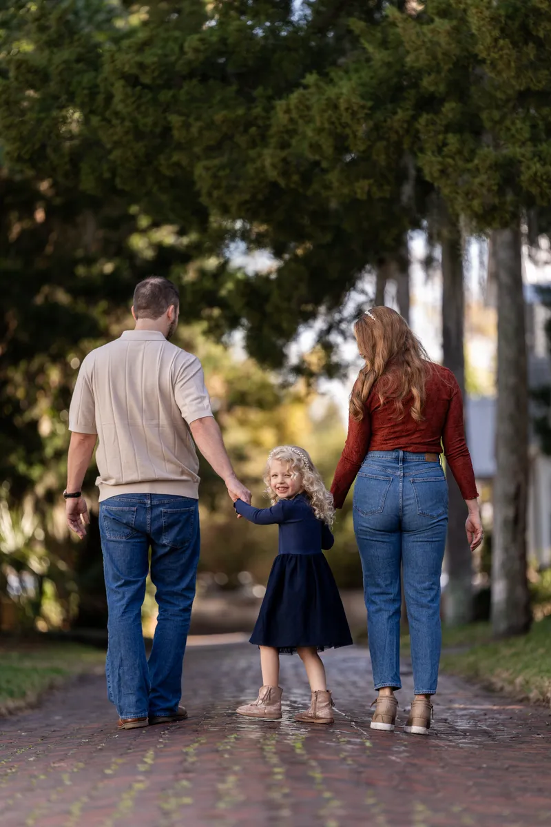 Ashley and Jordan walk hand-in-hand with their young daughter down a tree-lined brick pathway, creating a heartwarming family moment during their portrait session.