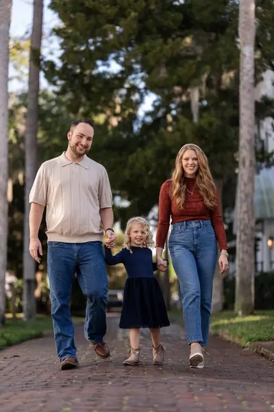 Ashley, Jordan, and their young daughter walk hand-in-hand down a tree-lined brick pathway, all smiling warmly at the camera in coordinated fall outfits.