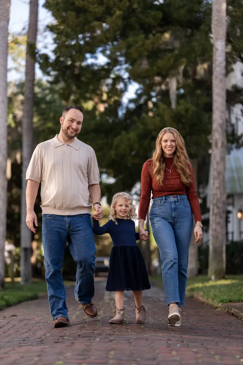 Ashley, Jordan, and their young daughter walk hand-in-hand down a tree-lined brick pathway, all smiling warmly at the camera in coordinated fall outfits.