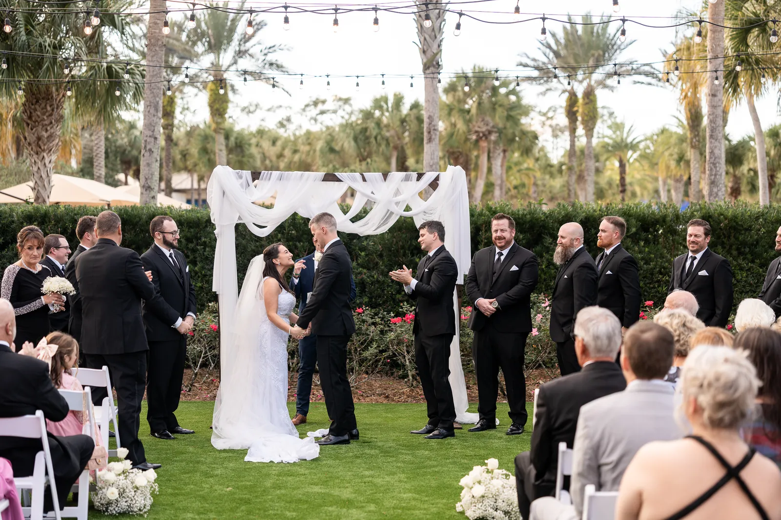 Jordan and Ashley stand together under an ivy-draped arch on the lawn, surrounded by seated guests and palm trees.
