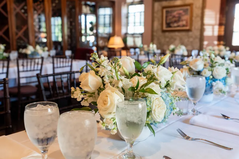 Close-up of a rehearsal dinner table setting with peach and white roses, greenery in a glass vase, water glasses, and white
