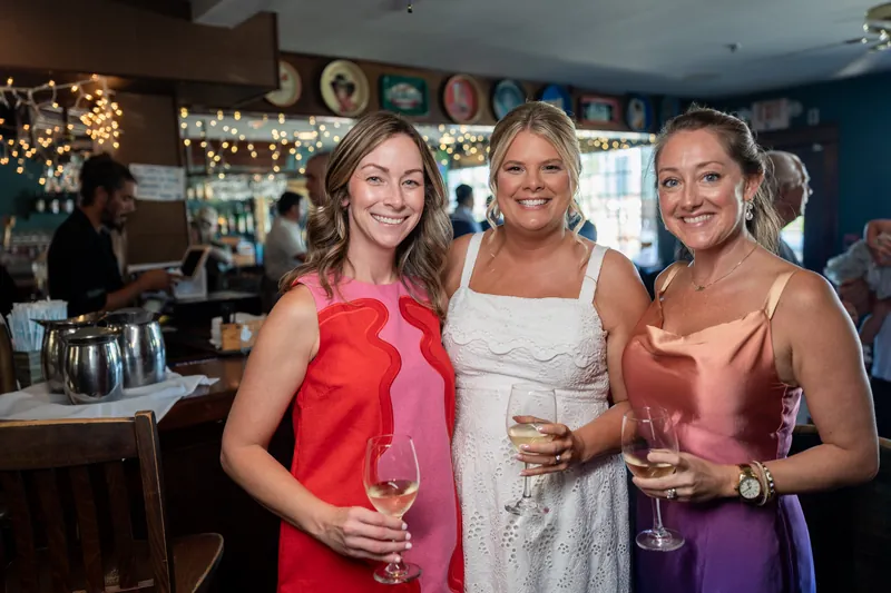 Three guests smile while holding wine glasses at the bar, with one in a red dress and another in white.