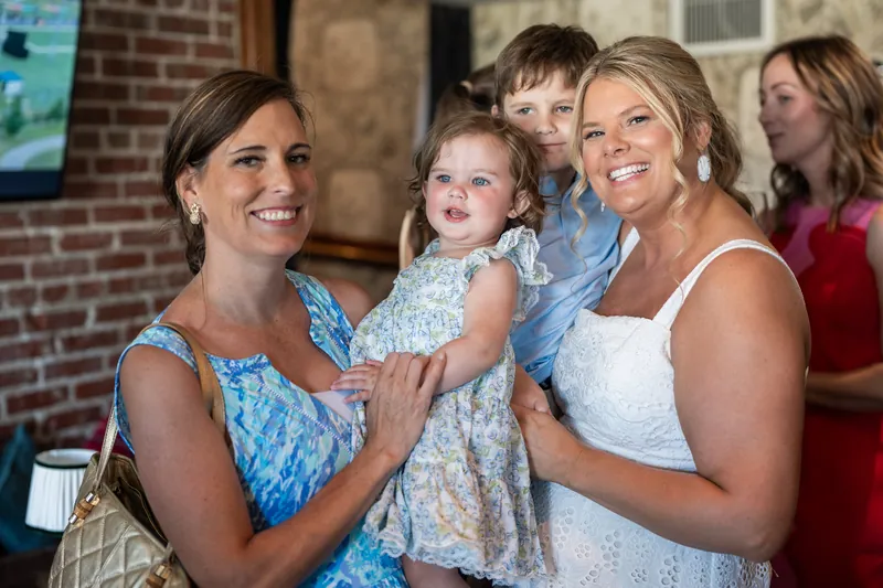 Two women and a young child pose together at the rehearsal dinner, all smiling at the camera near a brick wall.