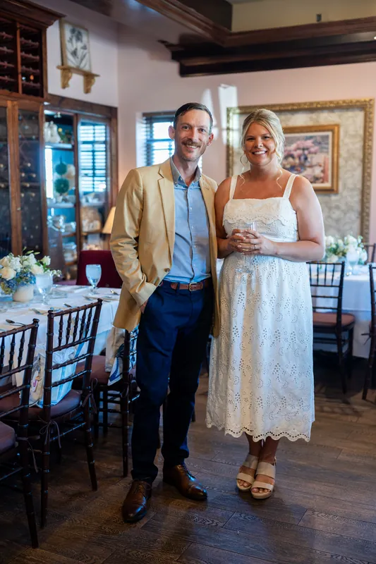 Jared wears a tan blazer and light blue shirt while Ashley stands beside him in a white lace dress, holding a drink.