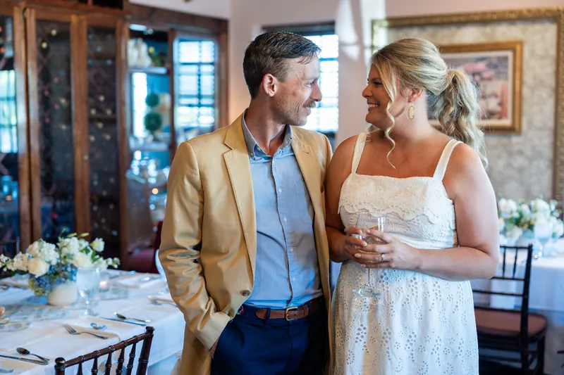 Ashley and Jared chat near a decorated table with white floral arrangements beneath a crystal chandelier.