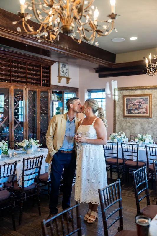 The couple stands together in the elegant dining space with dark wood cabinetry, framed artwork, and black Chiavari chairs