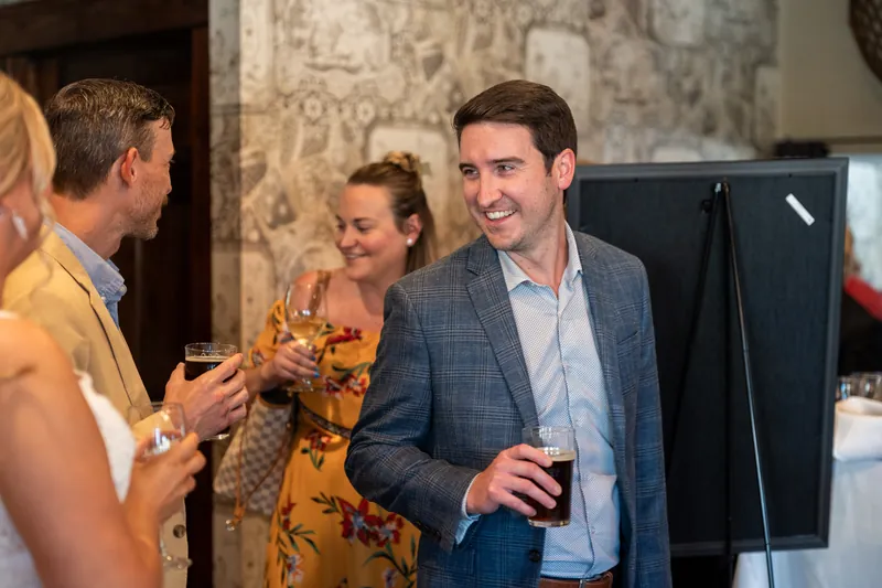 Jared smiles while holding a drink at The Chatsworth Pub and Tea Room as guests in yellow and light blue chat nearby.