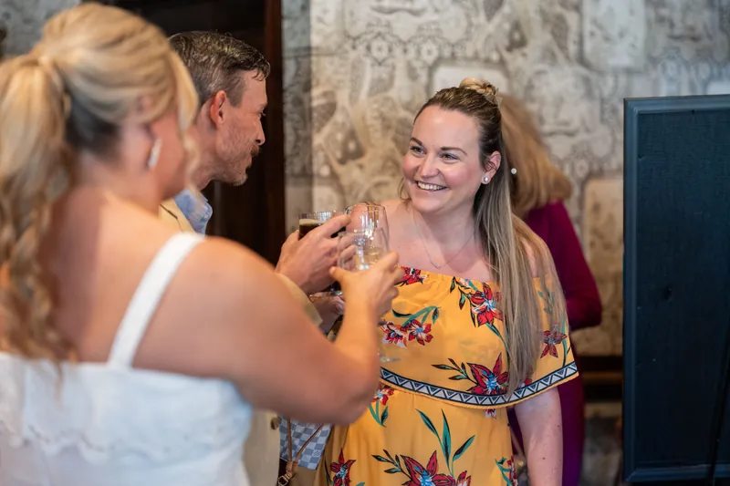 A woman in a yellow floral dress laughs while holding a glass, surrounded by friends at the rehearsal dinner.