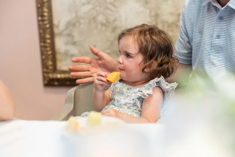 A young child in a floral dress eats at a white table while an adult watches nearby.
