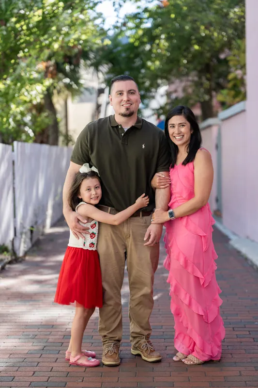 A man in olive and a woman in hot pink stand with two children on a sunny brick pathway lined with greenery.