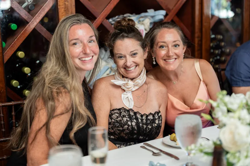 Three women smile at a dining table decorated with white flowers and glassware at the rehearsal dinner.