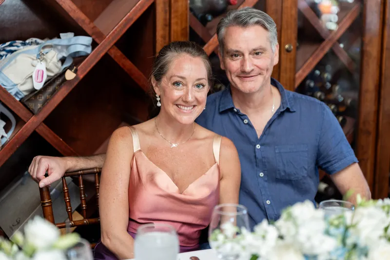 Ashley and Jared sit together at a white-draped table with white flower centerpieces and wine glasses between them.