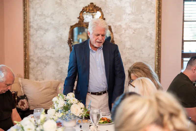 A man in a navy blazer stands to offer remarks during the rehearsal dinner as seated guests listen.