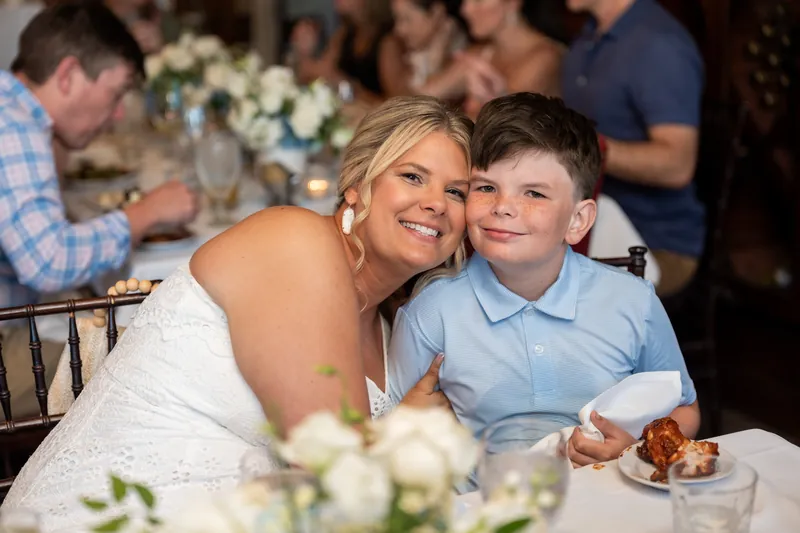 Ashley leans over to embrace a young boy in a light blue shirt at the dinner table, both smiling.