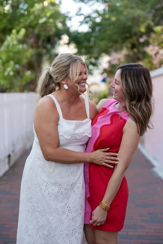 A woman in a white dress and a woman in a bright pink dress embrace outdoors on a brick path in Downtown St. Augustine.