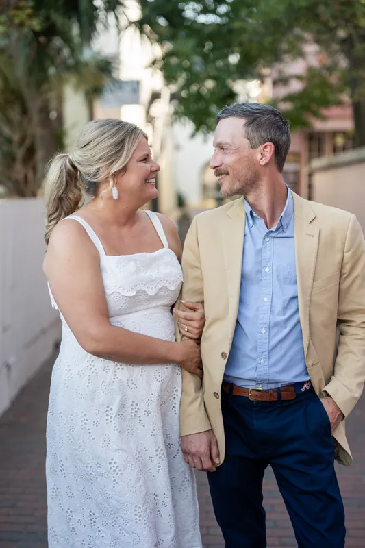 Ashley and Jared smile at each other while strolling through a tree-shaded Downtown St Augustine street, their arms linked.