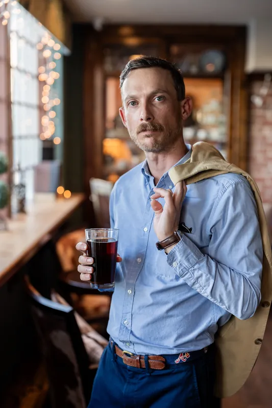 Jared stands at the bar in a light blue shirt, holding a dark drink.
