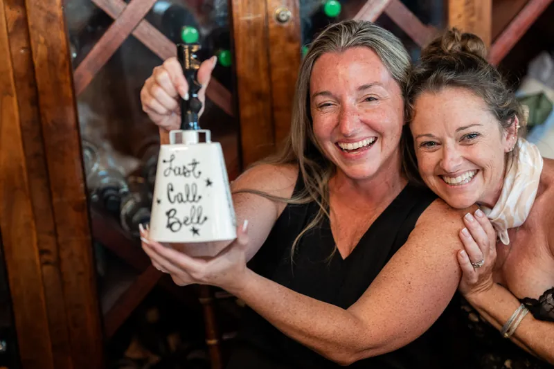Two women beam while holding a decorative bottle at The Chatsworth Pub and Tea Room.