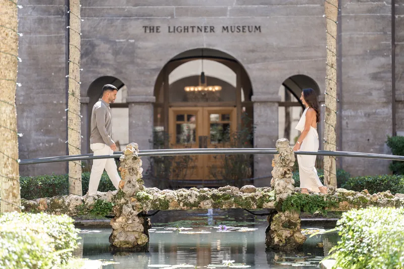 Two people in white stand on opposite sides of a reflecting pool, facing each other before the museum entrance.
