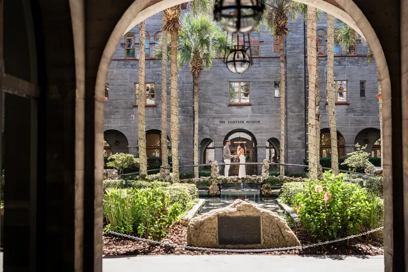 Stone archway frames a sunlit courtyard with tall palms, manicured hedges, and a person in white standing near a fountain at the Lightner Museum.