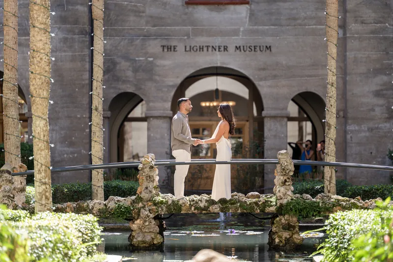 Person in white dress raises arms on a railed balcony overlooking a reflecting pool bordered by flowering plants and stone columns.