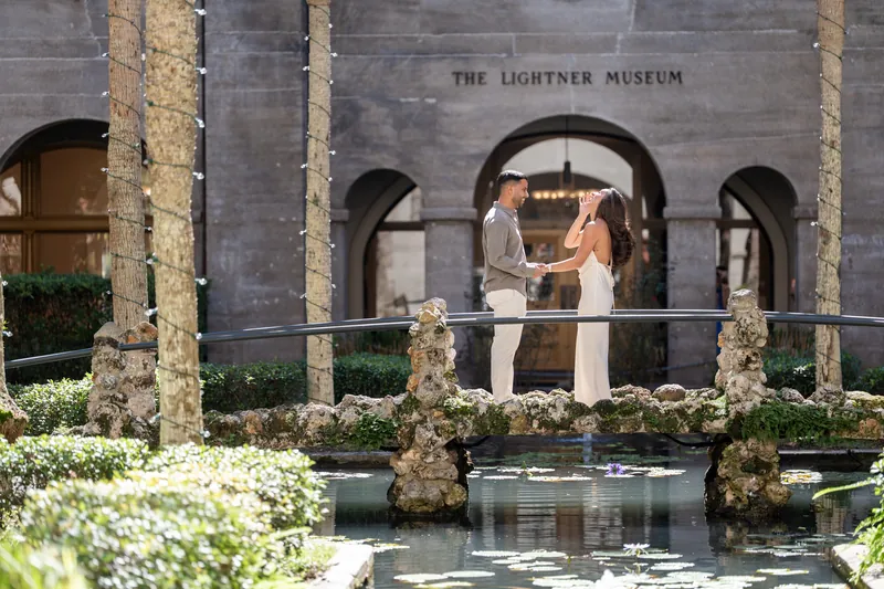 Two people stand facing each other on a balcony at the Lightner Museum, with arched columns and a reflecting pool below.