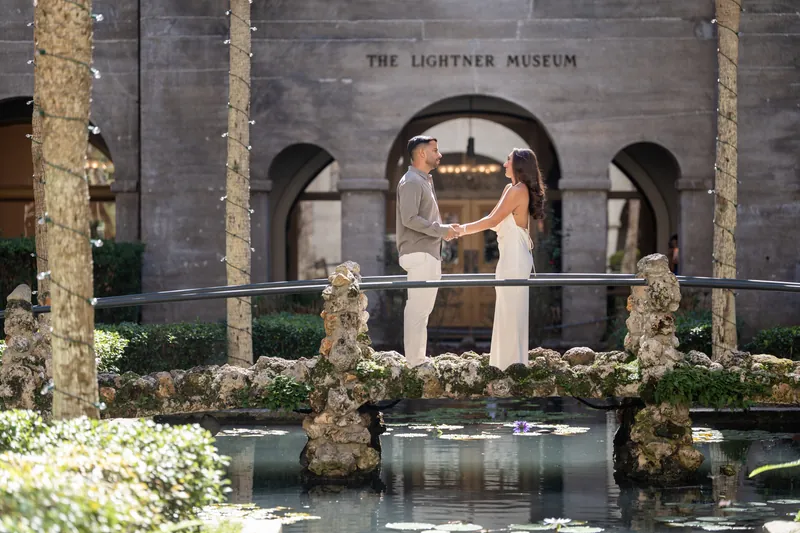 Two people hold hands on a garden balcony framed by moss-covered columns, the museum's stone archways visible across the reflecting pool.