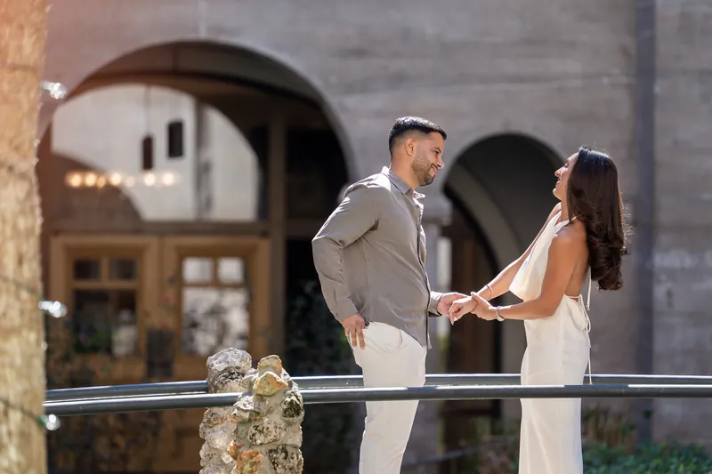 Close view of a couple in casual attire holding hands on a balcony, with arched stone architecture and warm interior lighting behind them.