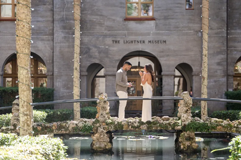 Couple stands together on a railed overlook above a reflecting pool, surrounded by flowering vines and the museum's classical columned courtyard.
