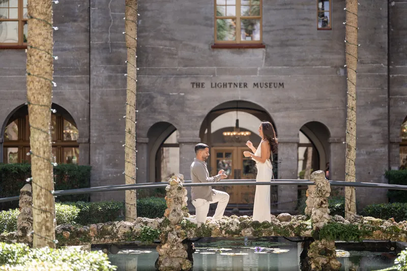 Two people stand on a central platform at the Lightner Museum surrounded by white flowers and tall palms against stone arches.