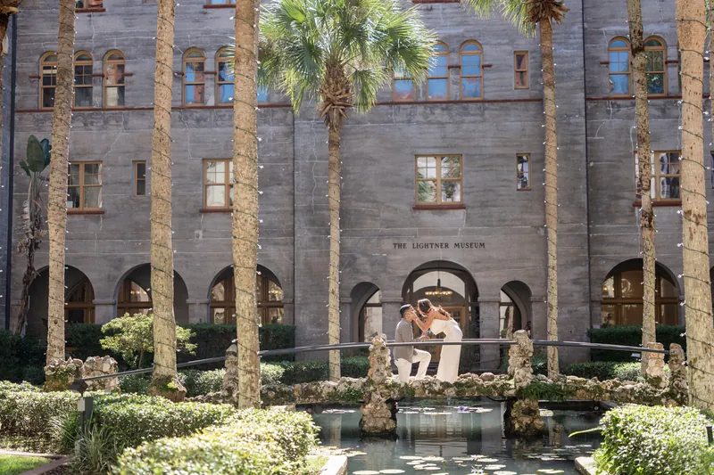 Two figures stand close on a decorated platform within the Lightner Museum's symmetrical courtyard lined with tall palms.