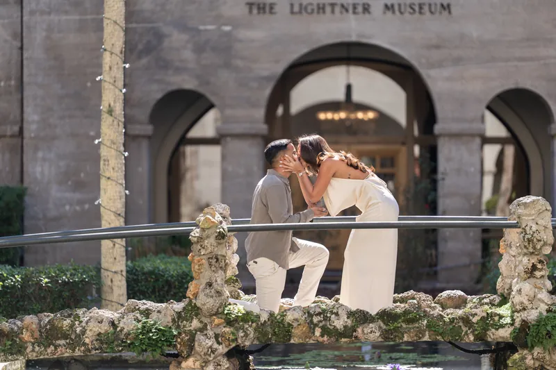 Two people in formal wear lean together on the museum platform, white floral garland draped along the railing behind them.