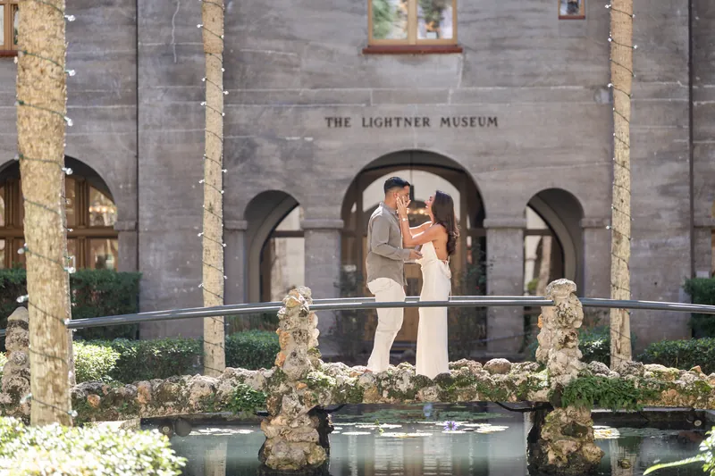 Two people embrace on a stone platform decorated with white flowers and greenery, arched columns rising on either side.