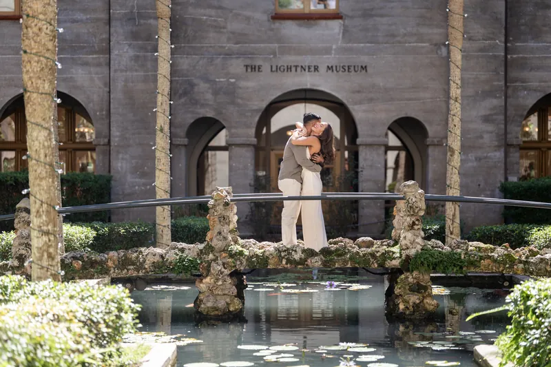 Two people stand on a moss-covered bridge at the Lightner Museum, with historic arches framing them above still water.