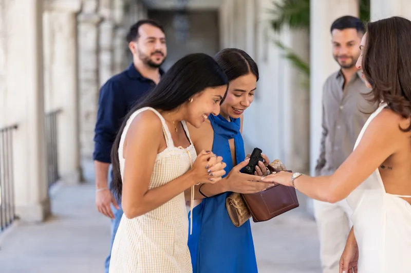 Someone in a white dress receives a small brown box from a friend while onlookers gather around in the courtyard.