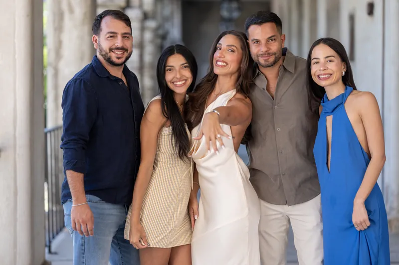 A group of five smiles at the camera in the covered walkway, with the person in the center wearing an elegant white wedding dress.