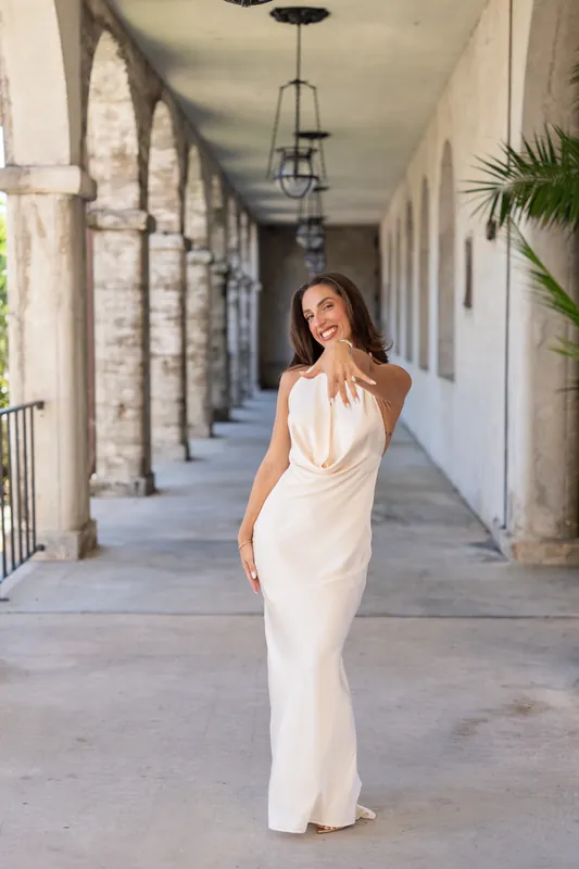 Someone in a cream-colored sleeveless gown walks alone down the colonnade, looking over their shoulder with a soft smile.