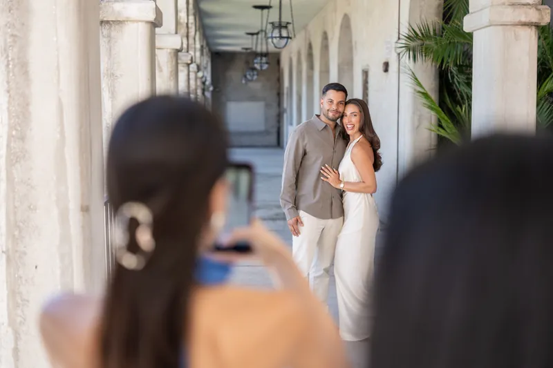 Two visitors photograph a couple posing together beneath the archway's pendant lights and white columns.