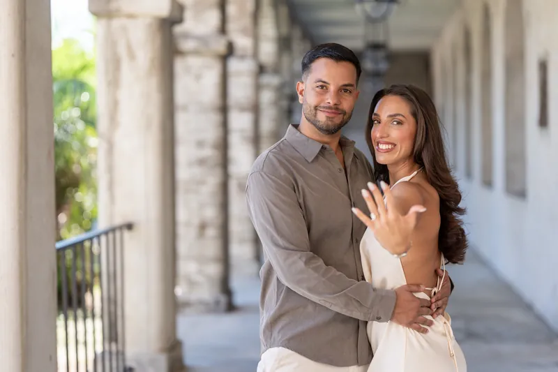 A couple embraces on the portico, with the person in the white gown displaying a ring on their left hand.