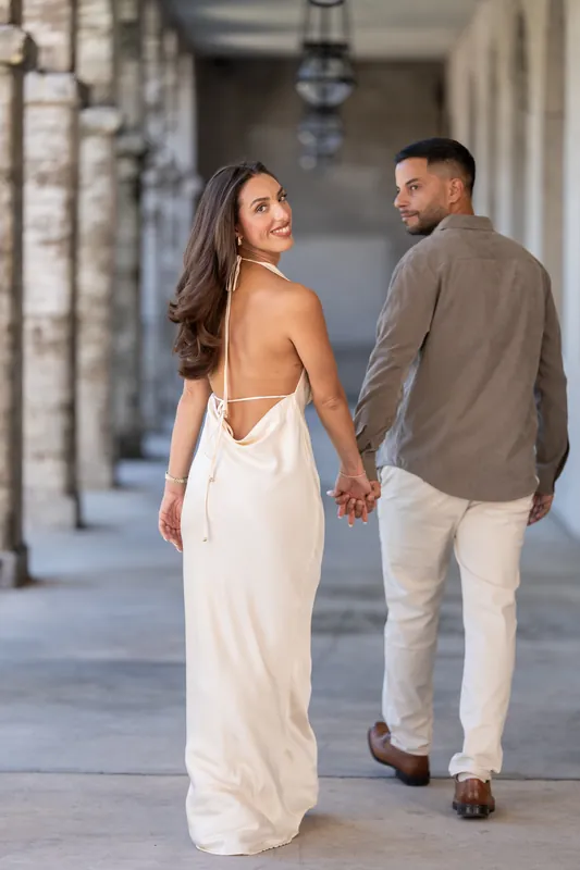 Two people hold hands while walking away down the colonnade, the one in white looking back toward the camera.