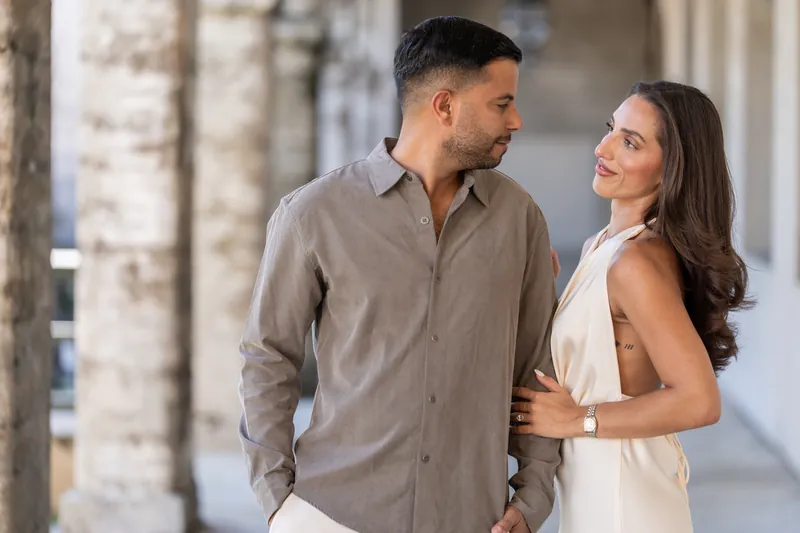 A couple pauses in the arcade's shade, the person on the left gazing at their partner in the white gown beside them.