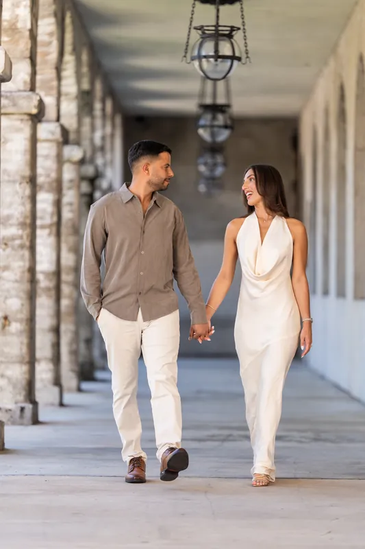 The couple walks toward the camera along the classical colonnade, their fingers intertwined as they move through dappled light.