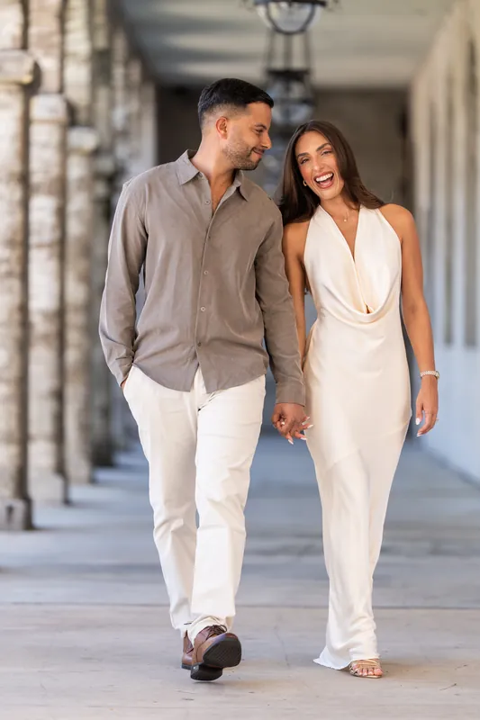 Beltran and Gabriela walk hand-in-hand through the Lightner Museum Courtyard's colonnade, he in a gray button-up and white pants, she in a flowing white sleeveless gown.