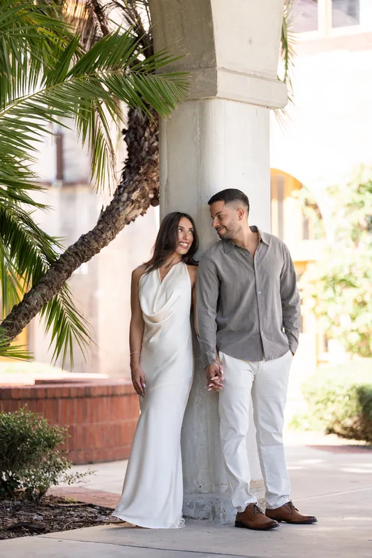 The two lean close together under palm fronds at the Lightner Museum, holding hands while gazing toward the sunlit courtyard behind them.