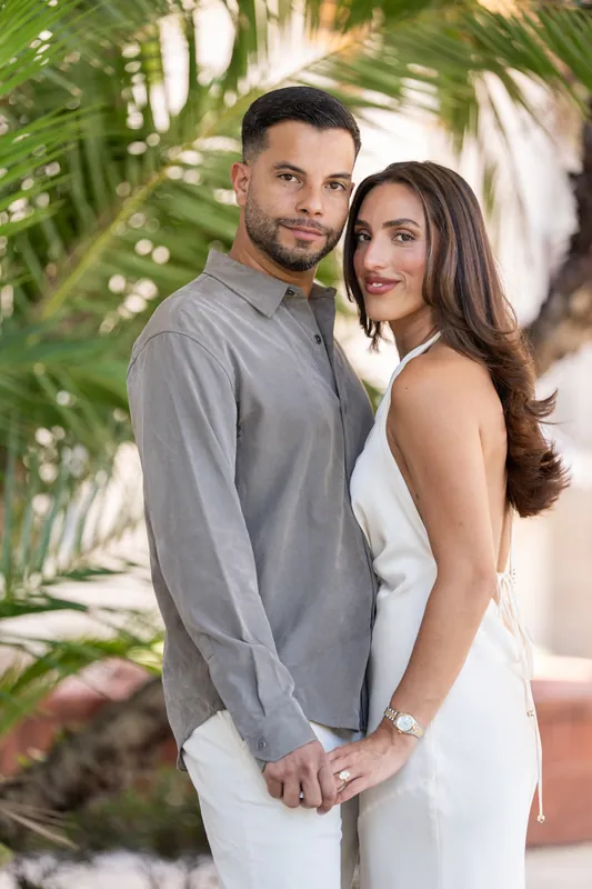 They stand chest-to-chest in dappled palm shade, foreheads nearly touching, both smiling with genuine warmth at the camera.